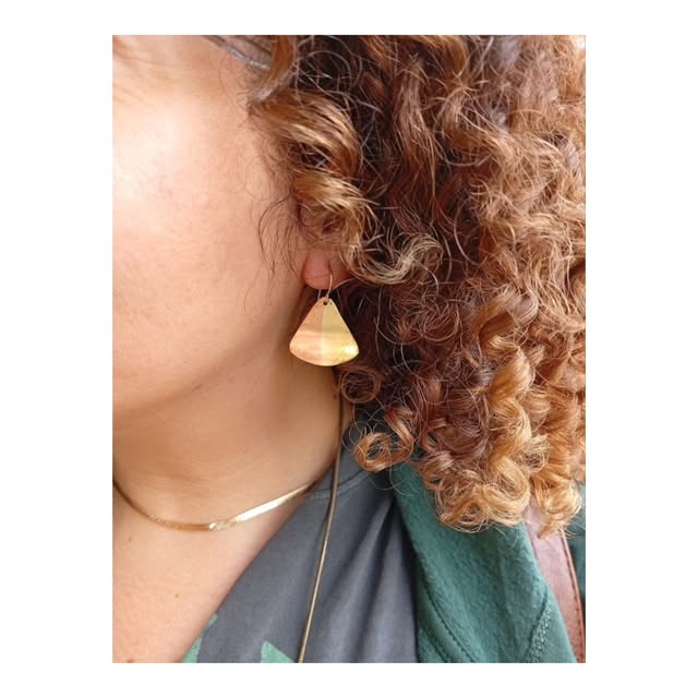 Gold-toned drop earring worn beside curly hair, with a slim silver-toned necklace.